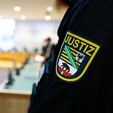 Gerichtssaal | Bildquelle: IMAGO, Jan Huebner Ärmel mit Logo eines Justizbeamten im Vordergrund, dahinter verschwommen der Gerichtssaal